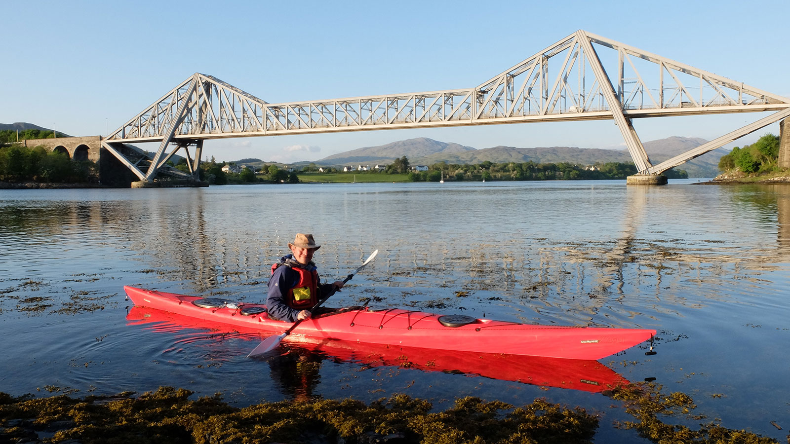 Grand Tours of Scottish Lochs. A man sits in a red cayak with a paddle. A bridge is behind him.